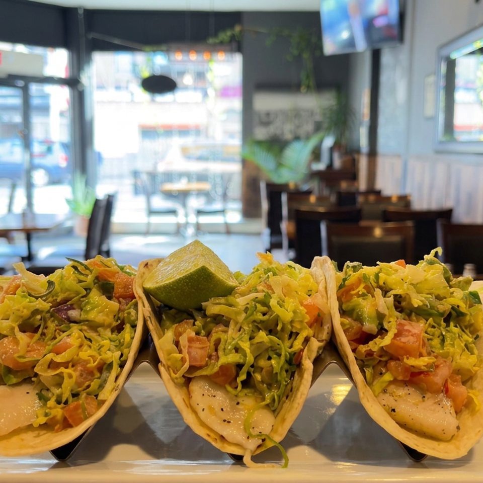 Three fish tacos topped with shredded lettuce, tomatoes, and lime, served on a plate in a bright restaurant dining room.